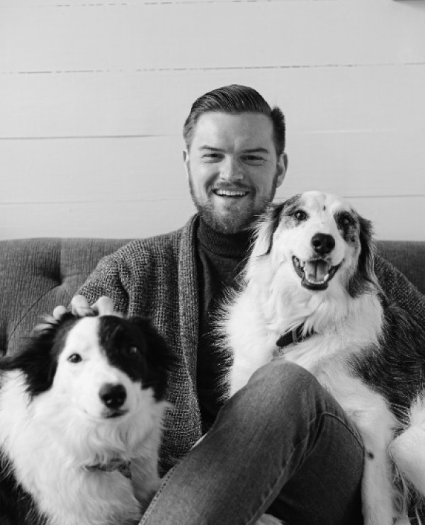 Smiling man in cozy sweater sits on couch with two joyful dogs, embodying warmth and companionship.