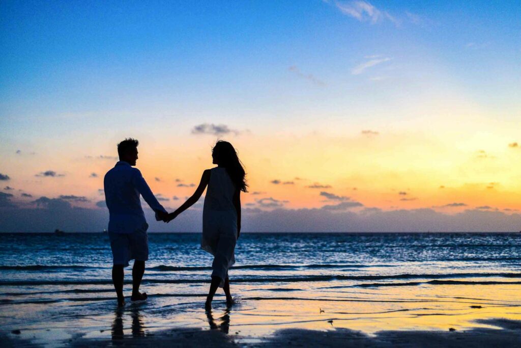 A romantic couple walks hand in hand on a tropical beach at sunset, enjoying a serene moment together.