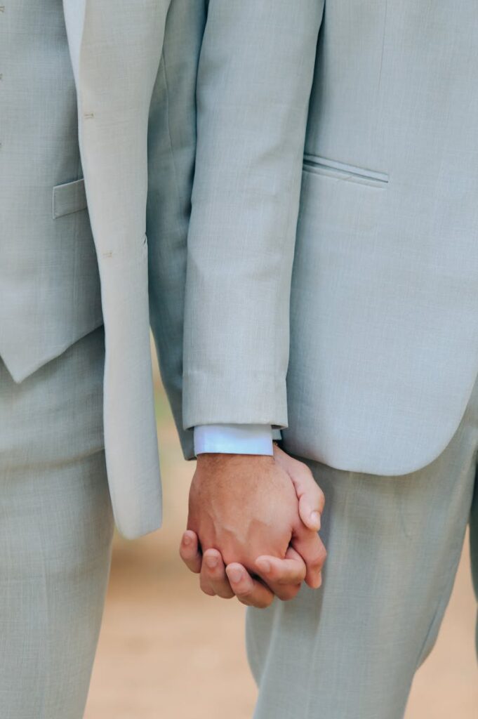 Intimate close-up of two men holding hands wearing pale suits, symbolizing love and unity.