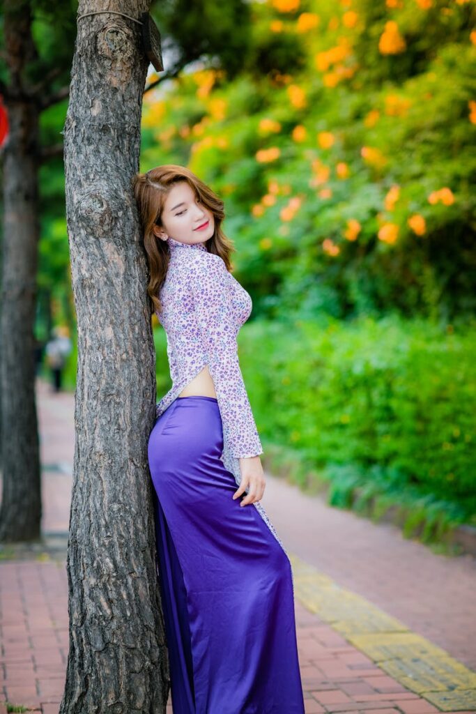 Femme élégante vêtue d'une robe violette, appuyée sur un arbre au feuillage éclatant. Parfait pour un éditorial de mode.