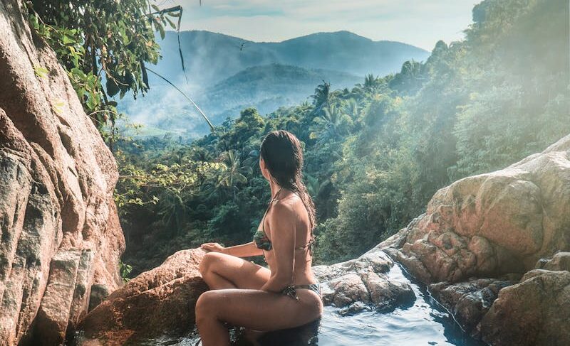 A woman in a swimsuit relaxes by a waterfall with lush mountains in Ko Samui, Thailand.