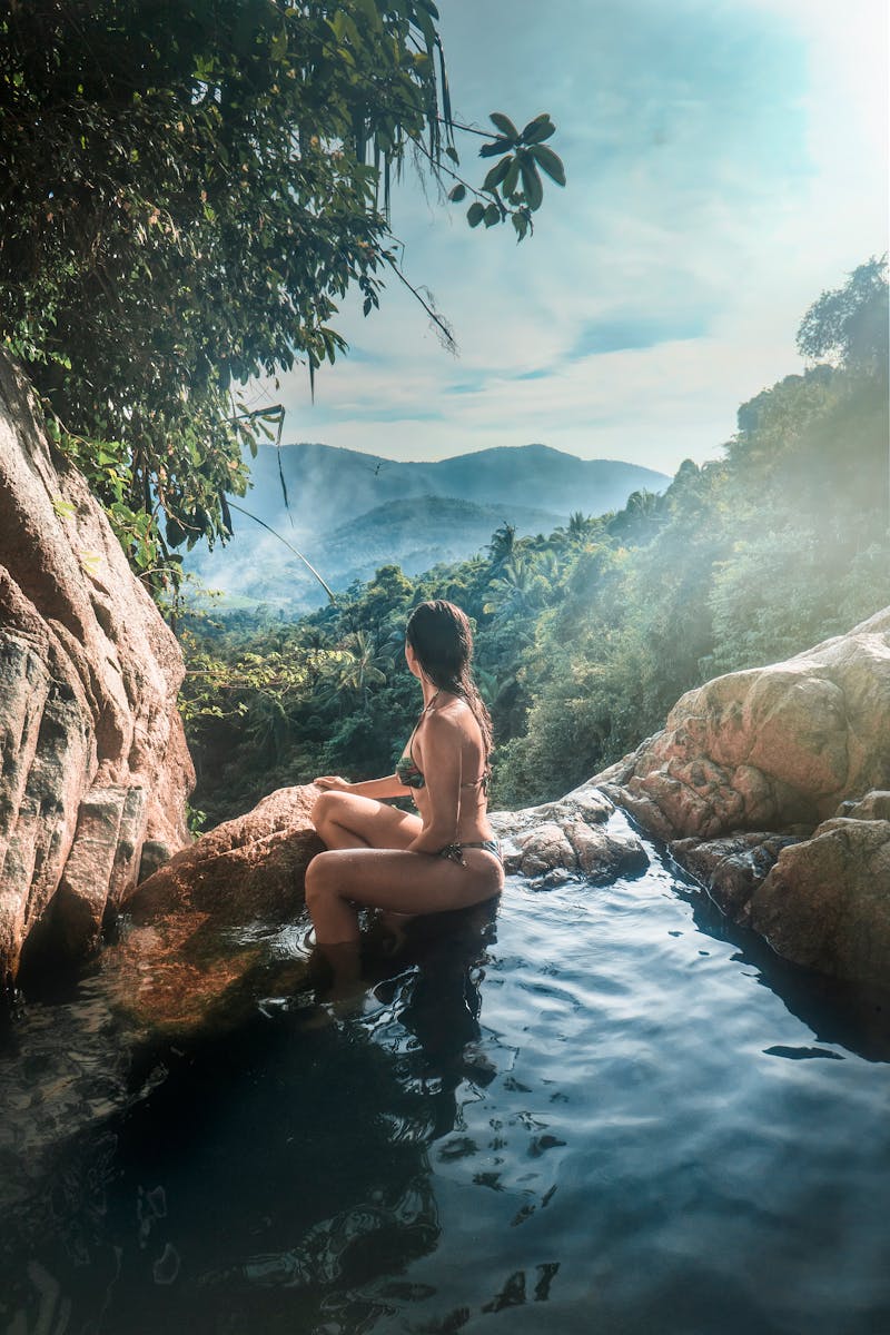 A woman in a swimsuit relaxes by a waterfall with lush mountains in Ko Samui, Thailand.