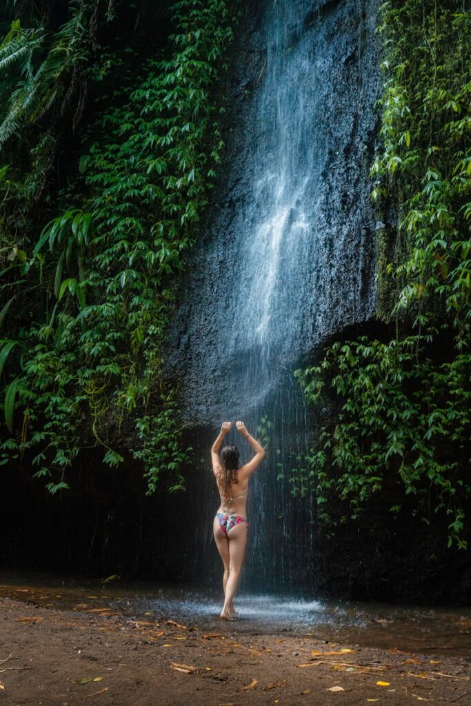 Back view of a woman enjoying a refreshing waterfall in a lush forest setting.