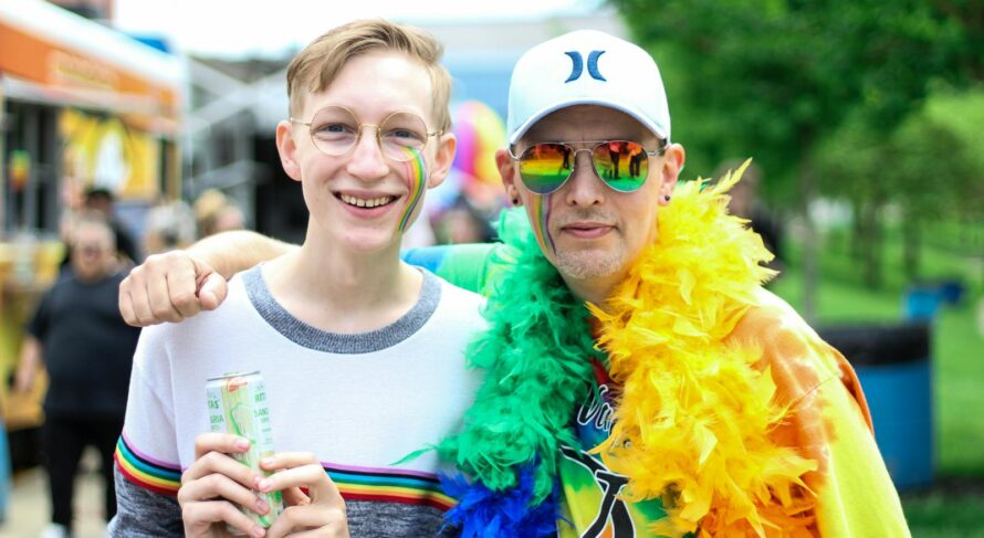 Two people smiling and embracing at a vibrant outdoor pride festival with rainbow colors.