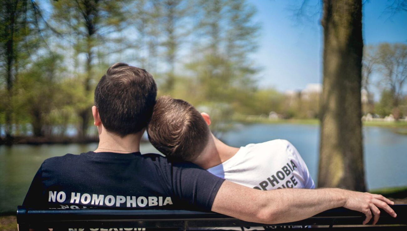 Two men sitting on a park bench by a lake, embracing love with unity and pride.