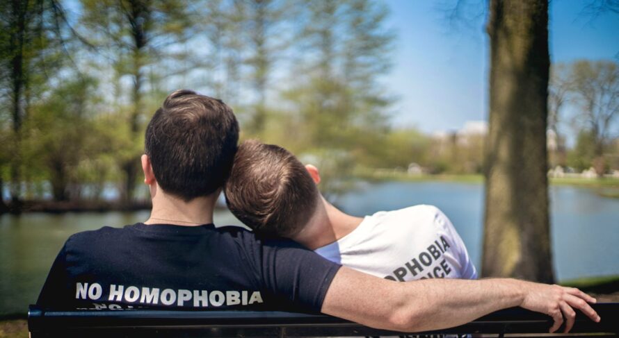 Two men sitting on a park bench by a lake, embracing love with unity and pride.