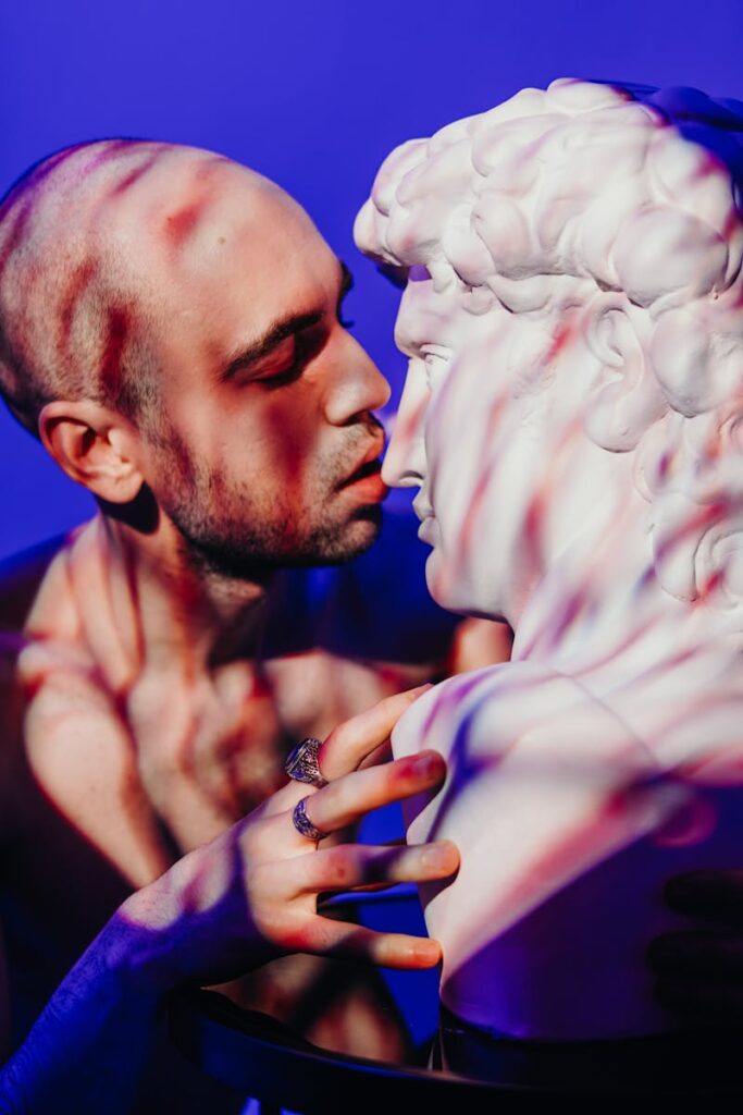 A bald, tattooed man intimately interacting with a classical sculpture under colorful light.