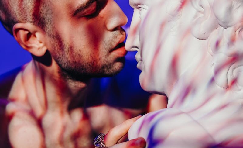 A bald, tattooed man intimately interacting with a classical sculpture under colorful light.