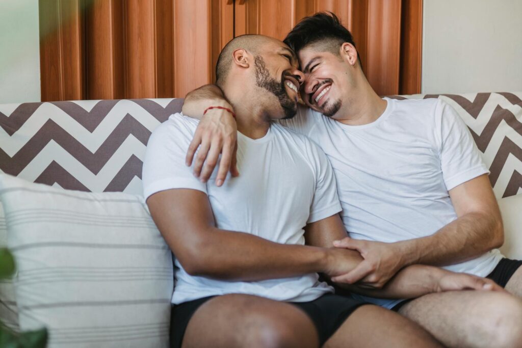 A joyful gay couple cuddling and smiling on a couch, showcasing affection and happiness.