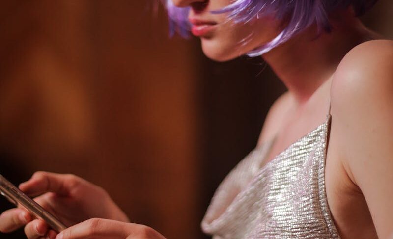 Young woman in violet wig and sparkly dress using a smartphone indoors.