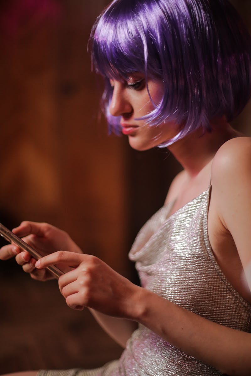Young woman in violet wig and sparkly dress using a smartphone indoors.