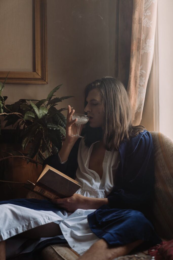 A contemplative man with long hair drinks wine and reads in a cozy indoor setting.