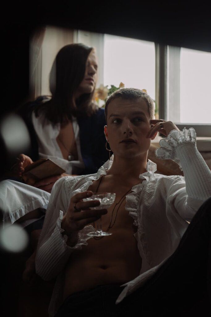 Two androgynous individuals lounge by a window, one holding a wine glass, exuding a refined and stylish aesthetic.