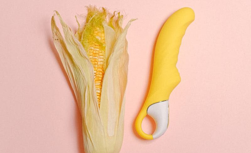 A creative still life featuring a corn cob and yellow silicone vibrator on a pastel background.