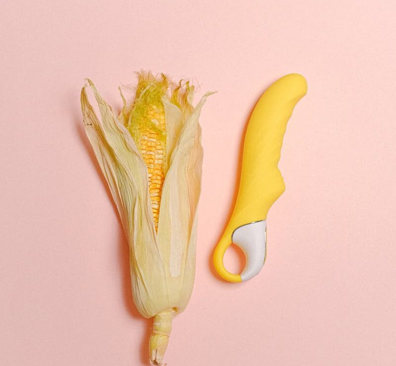 Nature morte créative présentant un épi de maïs et un vibrateur en silicone jaune sur un fond pastel.