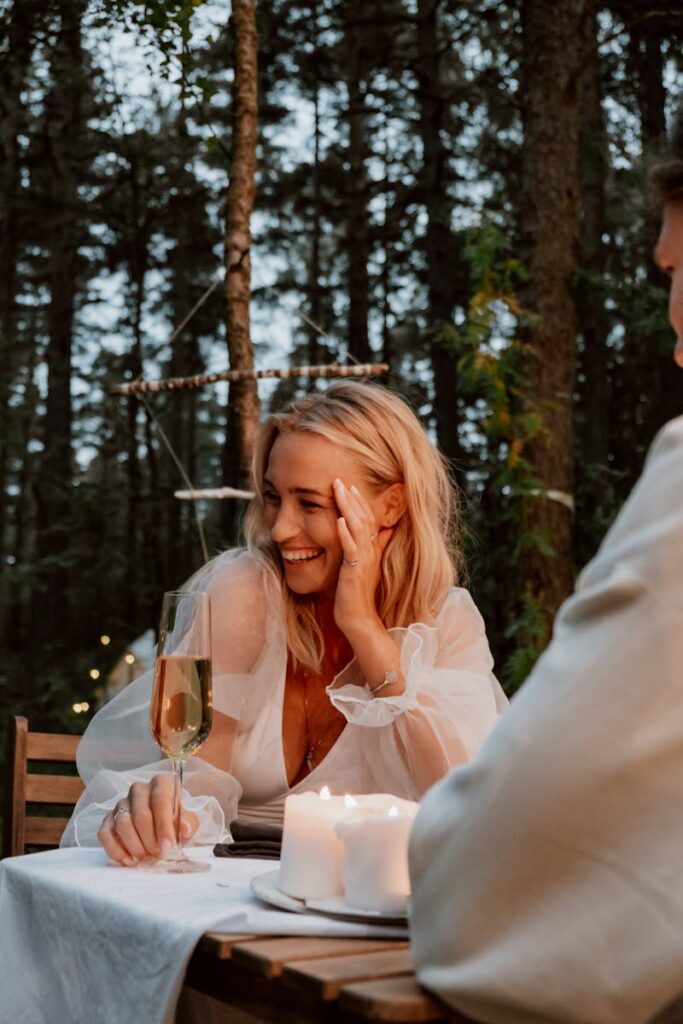 A woman enjoys a romantic outdoor dinner with champagne and candles in a serene forest setting.