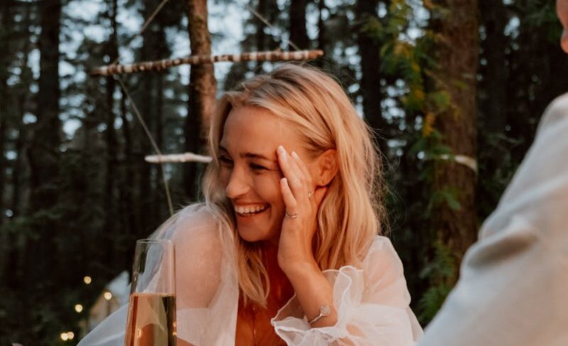 A woman enjoys a romantic outdoor dinner with champagne and candles in a serene forest setting.