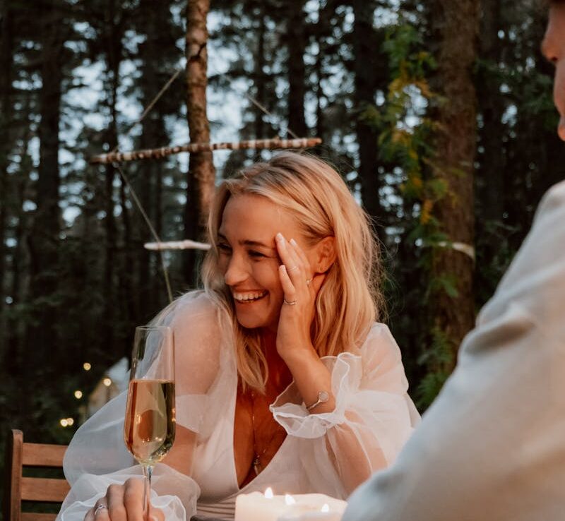 A woman enjoys a romantic outdoor dinner with champagne and candles in a serene forest setting.