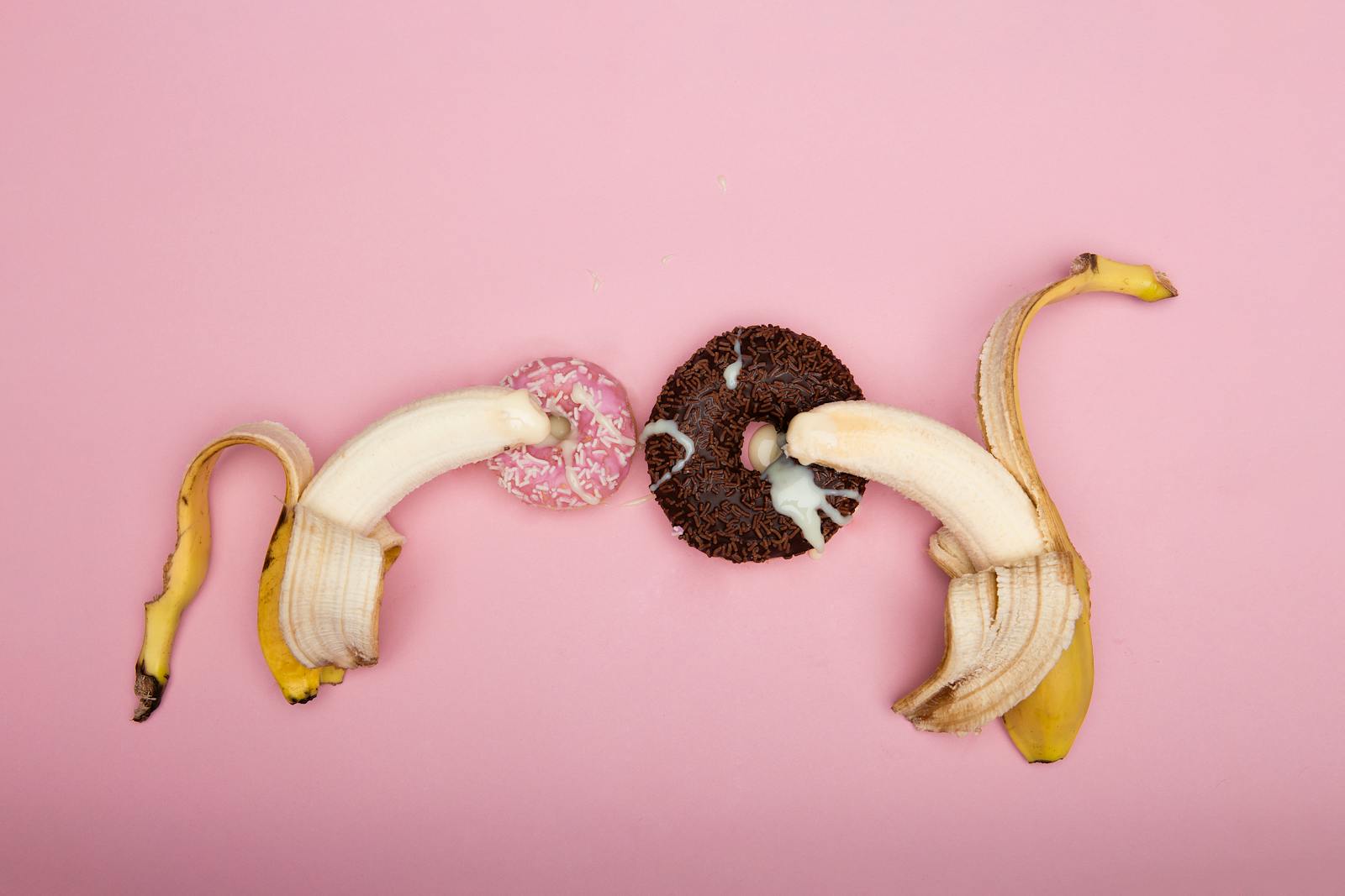 Disposizione giocosa di banane sbucciate e ciambelle su sfondo rosa, perfetta per l'arte concettuale.