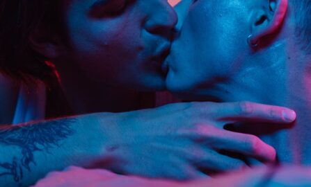 Amanti tatuati che si scambiano un bacio emotivo Un tenero momento catturato mentre due uomini si scambiano un bacio sotto una vibrante illuminazione al neon, simbolo di amore e inclusione.