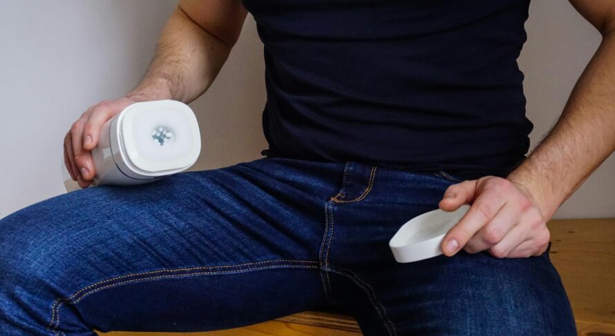 A man seated indoors holding an adult toy device, wearing jeans and a black shirt, with focus on the product.