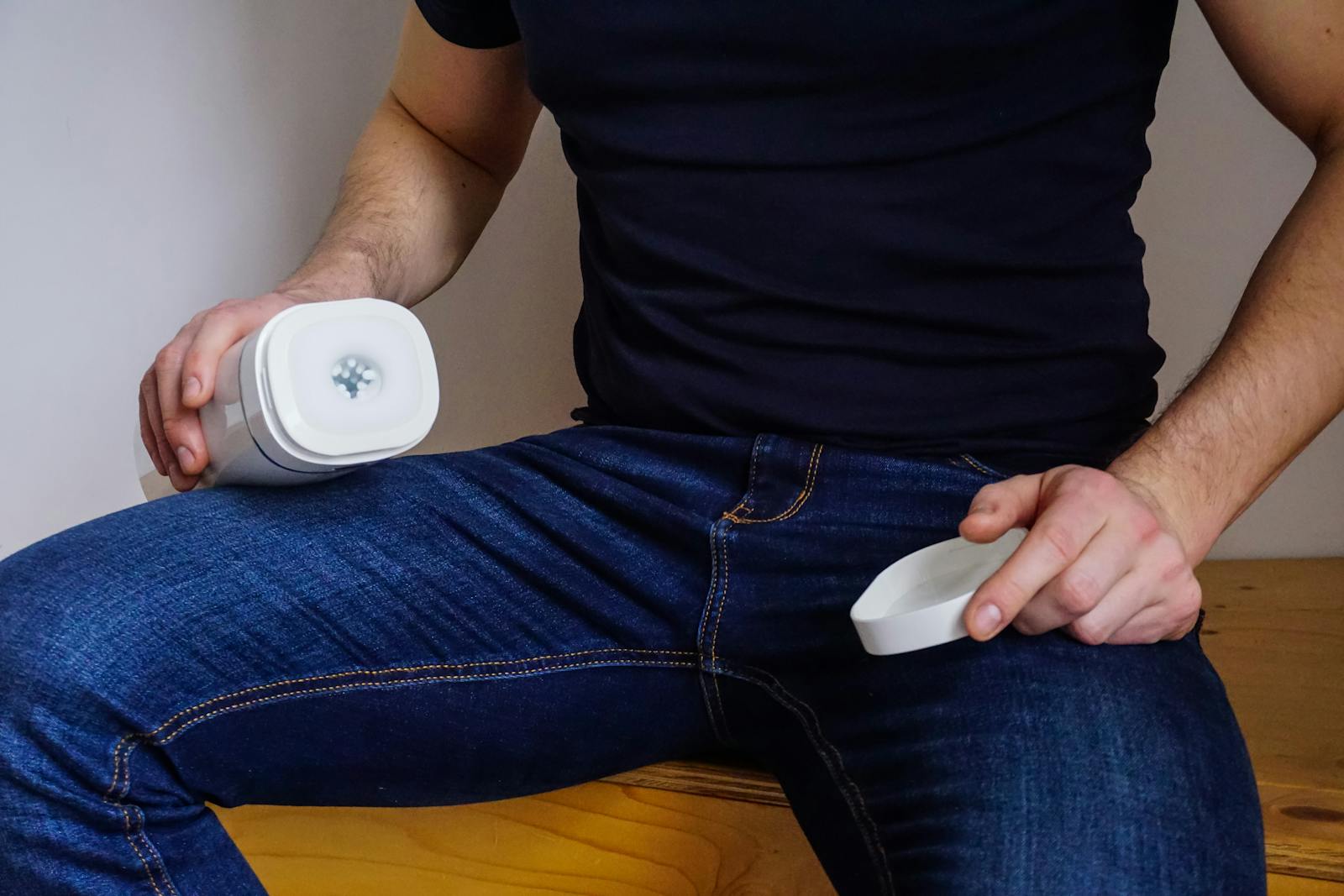 Uomo seduto in casa con in mano un dispositivo giocattolo per adulti, in jeans e camicia nera, con focus sul prodotto.