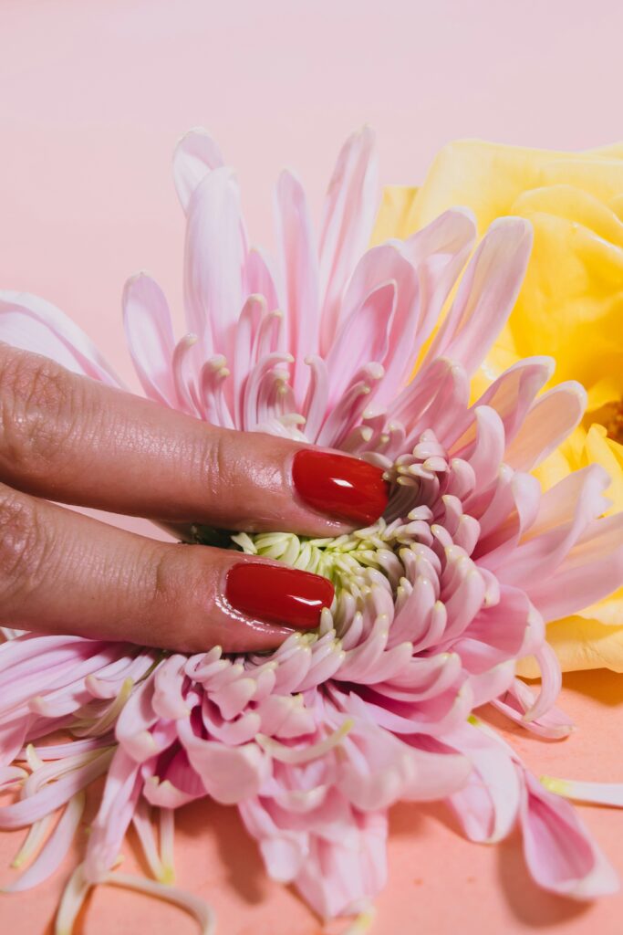 Mão com unhas vermelhas por cima de um ramo de flores cor-de-rosa sobre fundo pastel.