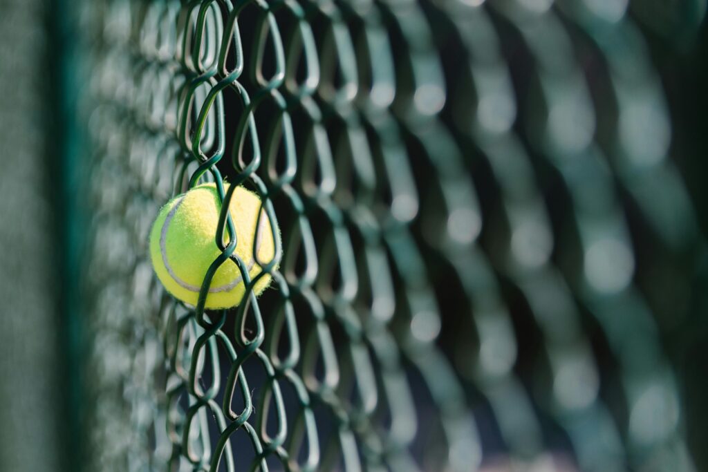 Tennis ball caught in green chain-link fence, symbolizing unexpected moments in sports.