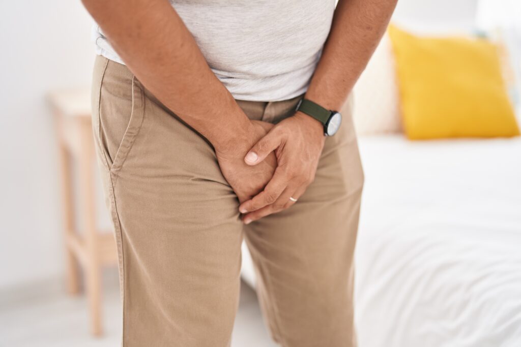 Young man in bedroom showing discomfort while covering his genitals with hands.