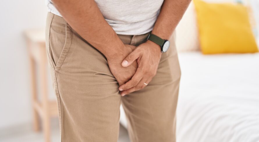 Young man in bedroom showing discomfort while covering his genitals with hands.