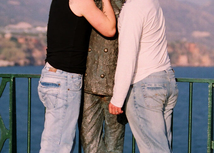 Two men share a playful moment with a statue in a picturesque coastal setting, highlighting friendship and humor in a vibrant landscape.