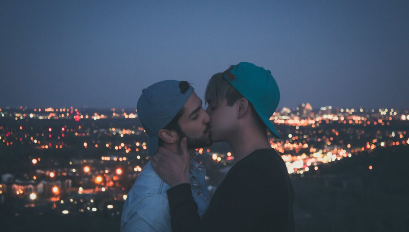 A joyful gay couple shares a kiss under the mesmerizing city lights at twilight, embracing their love in a vibrant urban setting.