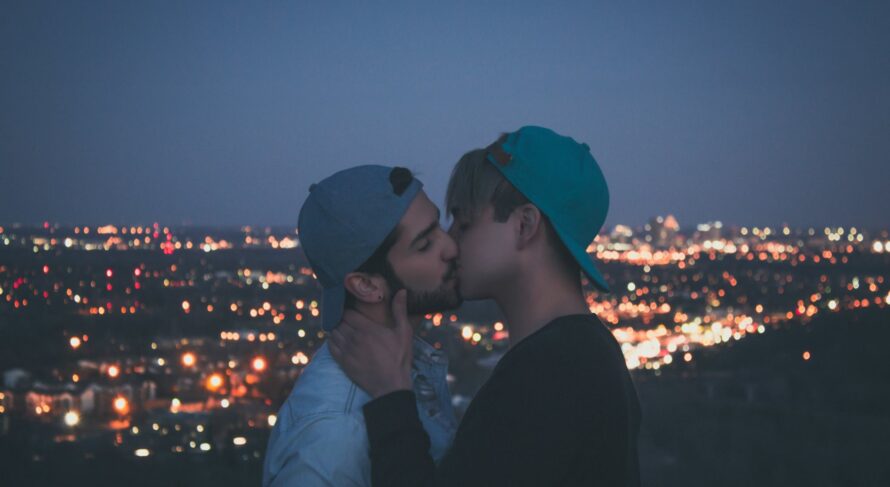 A joyful gay couple shares a kiss under the mesmerizing city lights at twilight, embracing their love in a vibrant urban setting.